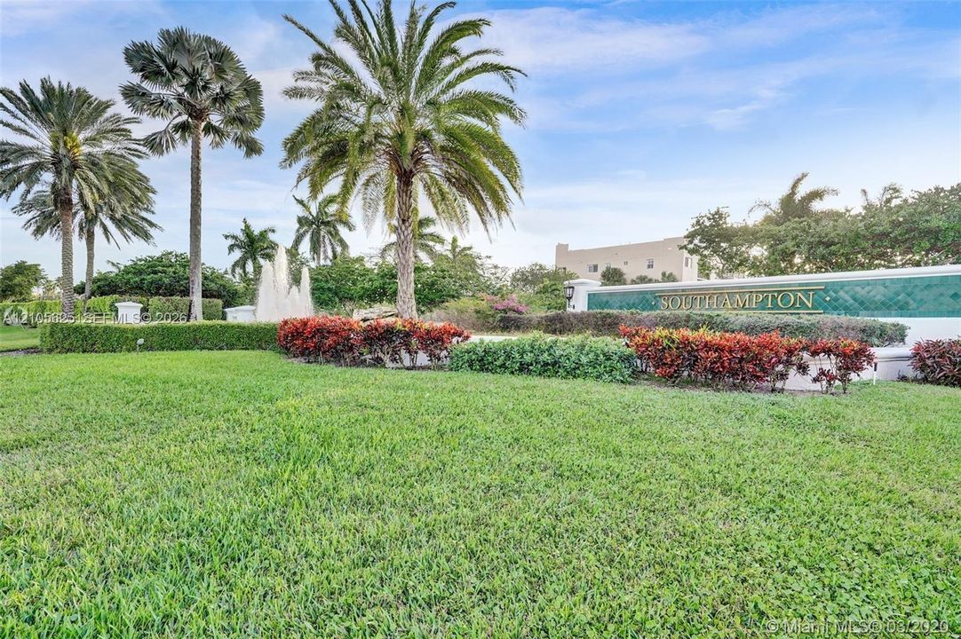 Community entrance featuring manicured green space, mature palm trees, decorative shrubbery, and a stone water fountain