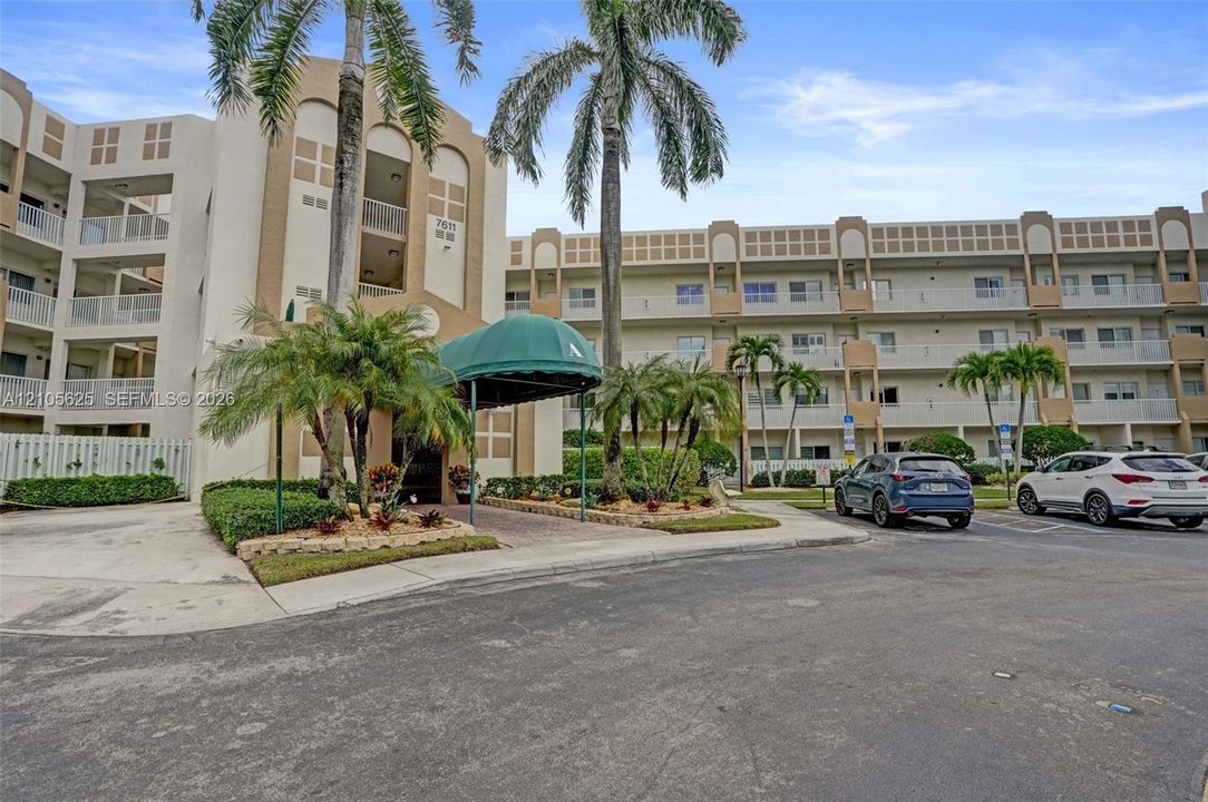 Multi-story residential complex featuring stucco exterior facade, green arched awning entry, paved walkway, and mature palm trees