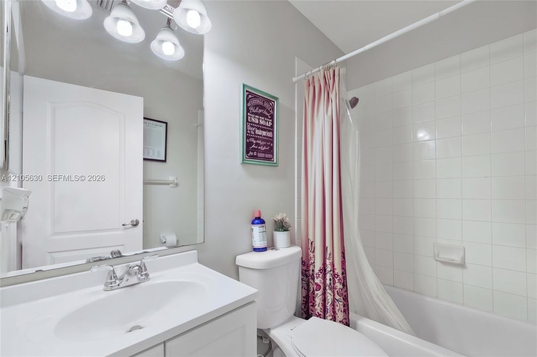 Vanity featuring an integrated sink with a chrome faucet, large frameless wall-mounted mirror, four-bulb overhead vanity lighting, and a white tiled shower surround