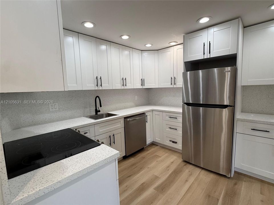 Kitchen featuring white Shaker-style cabinetry, light-toned composite countertops, stainless steel appliances, matte black hardware, and wood-finish flooring