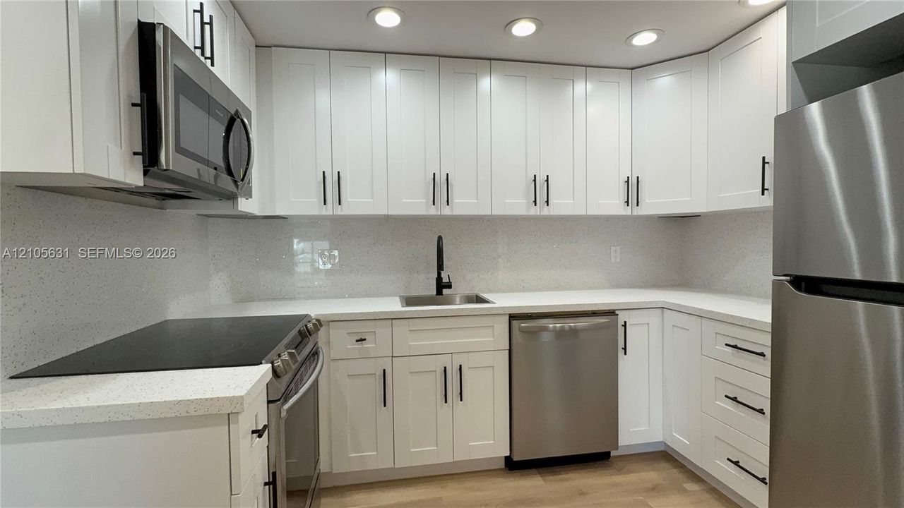 Kitchen featuring white Shaker-style cabinetry, light-toned composite countertops, a full-height solid surface backsplash, stainless steel appliances, and matte black hardware