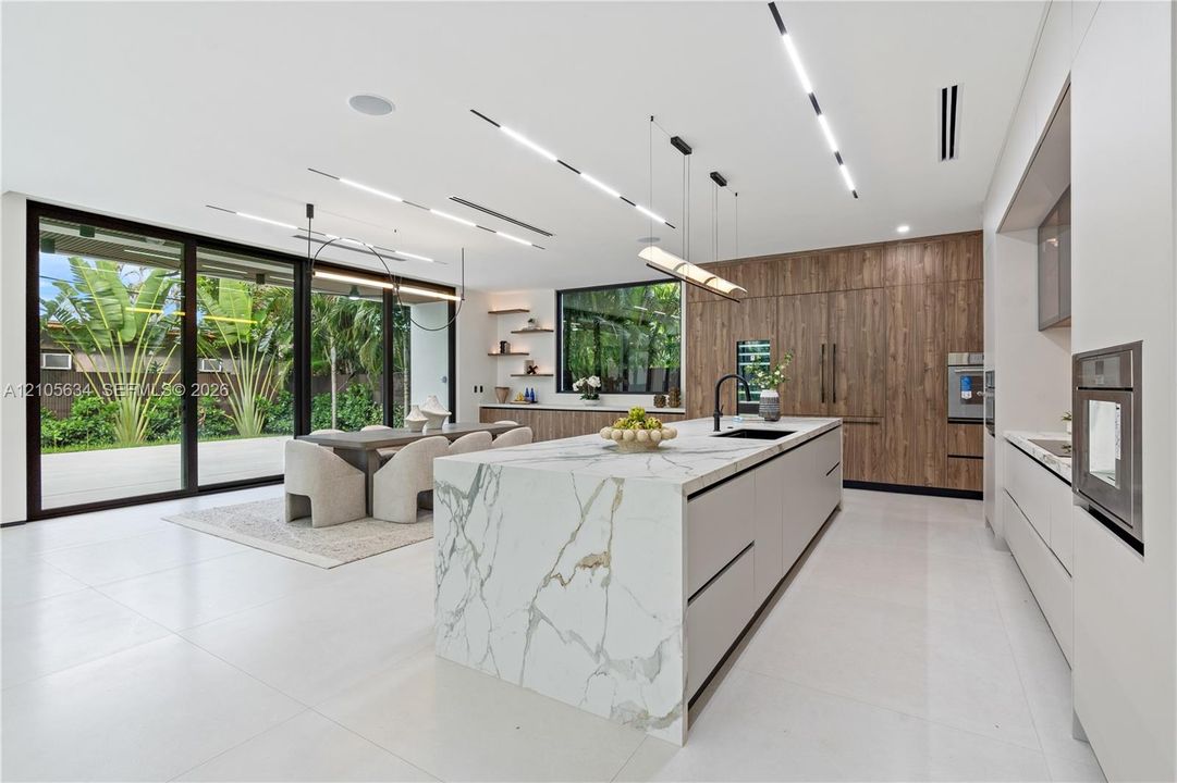 Kitchen featuring an oversized island with stone-finish waterfall countertop, wood-finish cabinetry, floor-to-ceiling glass sliding doors, integrated linear lighting, and large-format tile flooring