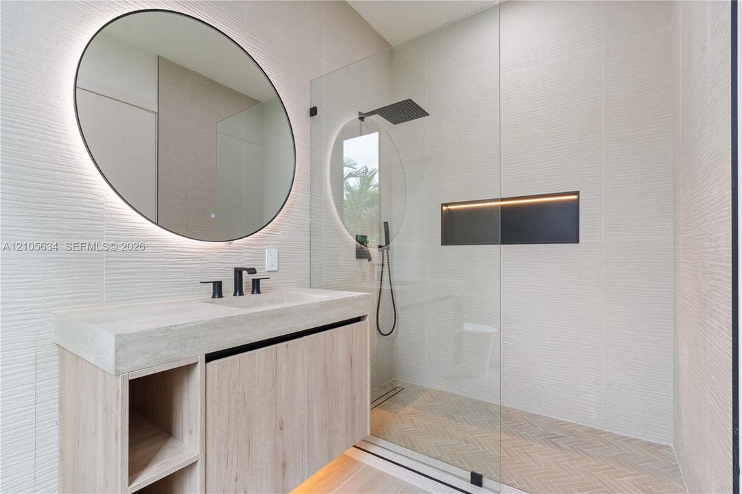 Floating vanity with integrated sink, textured wall tile, circular backlit mirror, frameless glass shower enclosure with rainfall showerhead, and herringbone-patterned shower floor tile