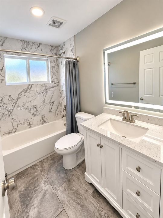 White cabinetry with stone-finish countertop, undermount sink, LED-backlit mirror, patterned stone-finish shower surround, and large-format tile flooring