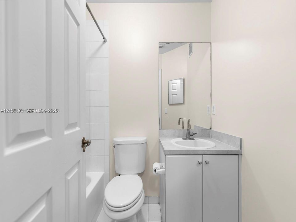 Bathroom featuring a vanity with a solid-surface countertop, an integrated sink, a wall-mounted mirror, a tub-shower combination with white tile surround, and a low-flow toilet