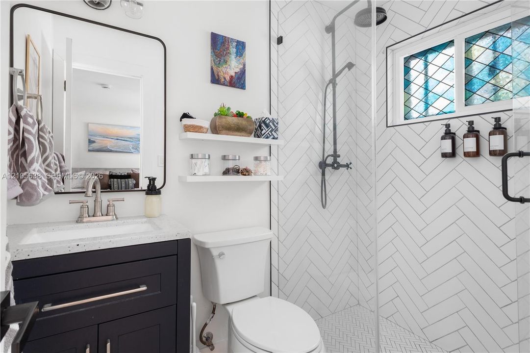 Bathroom featuring a dark-finish vanity with a speckled countertop, a walk-in shower with white herringbone-patterned wall tile, a glass shower enclosure, and a window with decorative leaded-glass detailing