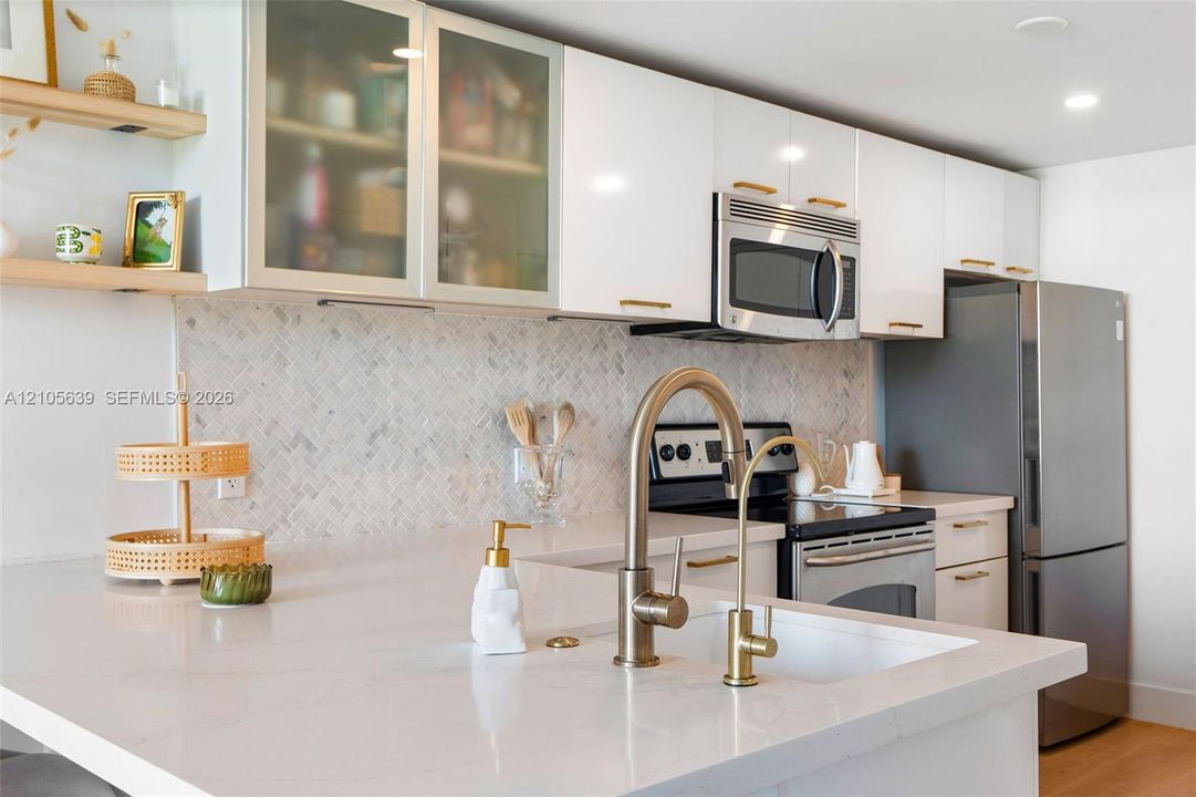 Kitchen featuring white cabinetry with glass-front uppers, herringbone-patterned stone backsplash, quartz countertops, brass-finish fixtures, and built-in stainless steel appliances