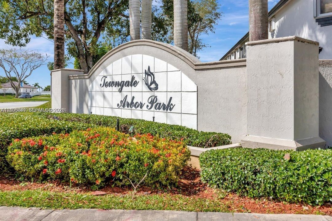 Community signage featuring stucco masonry walls, tile paneling, and manicured landscaping with blooming shrubbery