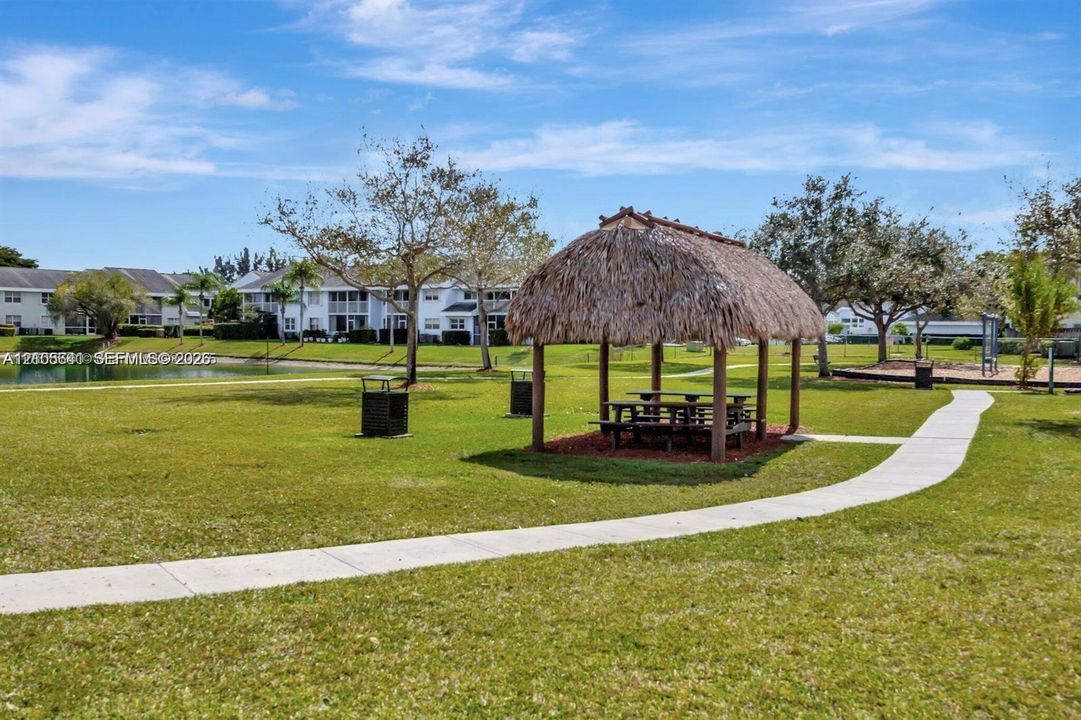 Outdoor pavilion featuring a thatched roof, expansive lawn, mature trees, winding concrete pathway, and waterfront proximity