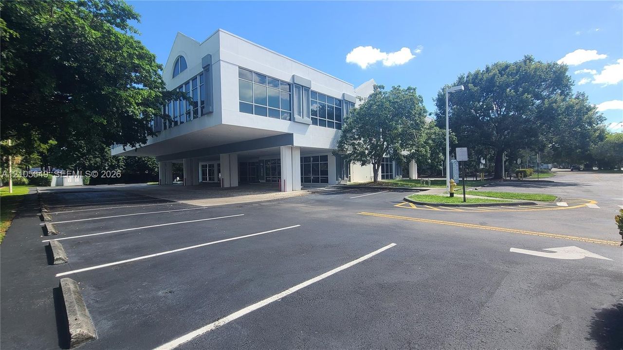 Multi-story professional building featuring a cantilevered exterior structure, expansive glass windows, paved surface parking, and mature landscape elements