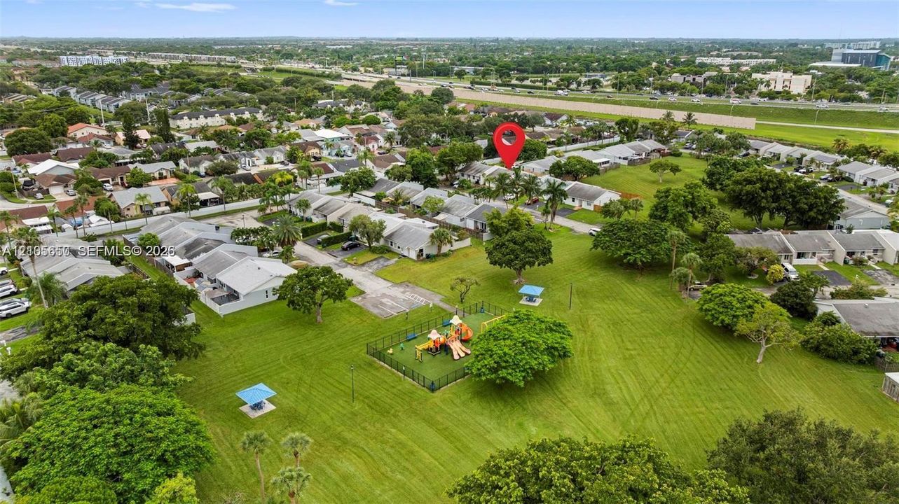 Aerial perspective of residential area featuring expansive green space, mature canopy trees, paved pathways, and a fenced playground structure