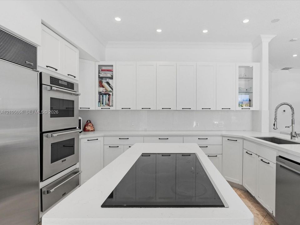 Kitchen featuring white cabinetry with glass-front display panels, quartz countertops, a central island with an integrated cooktop, double wall ovens, and a gooseneck faucet