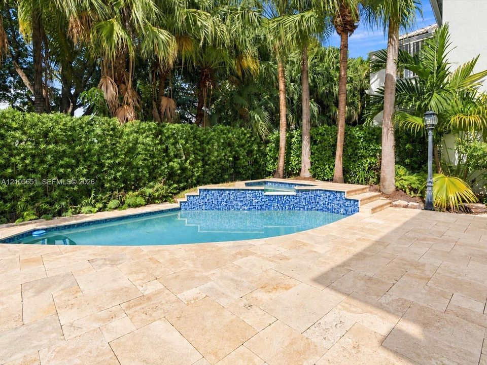 Outdoor area featuring a stone-paver patio, a swimming pool with blue tile accents, a raised spillway, and mature perimeter landscaping