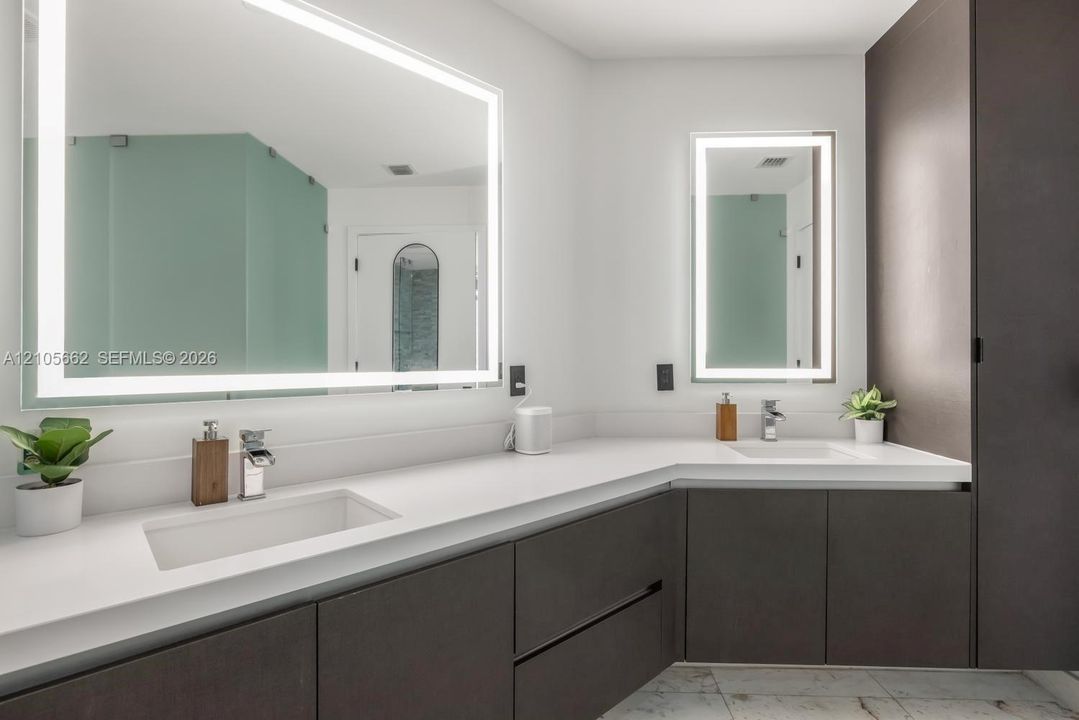 Double vanity featuring dark cabinetry, solid-surface countertops, integrated rectangular sinks, polished chrome faucets, and large backlit mirrors
