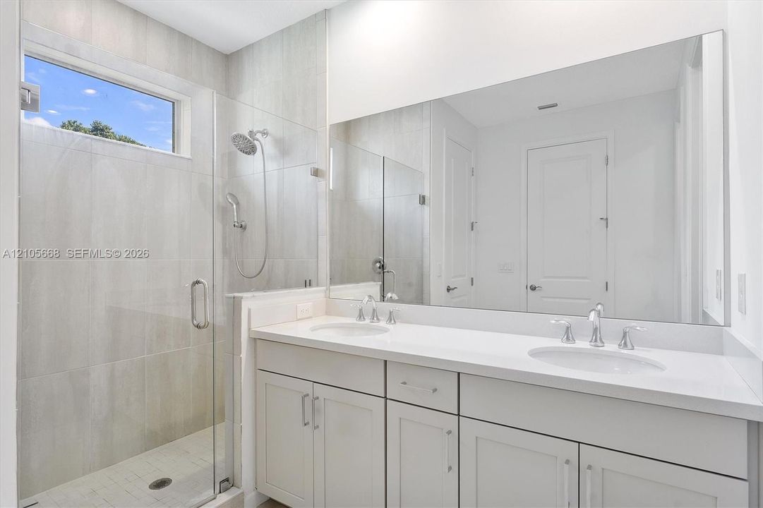 Double vanity featuring white cabinetry, solid surface countertops, dual undermount sinks, and a full-width wall mirror, alongside a glass-enclosed shower with tile surround and adjustable handheld shower head