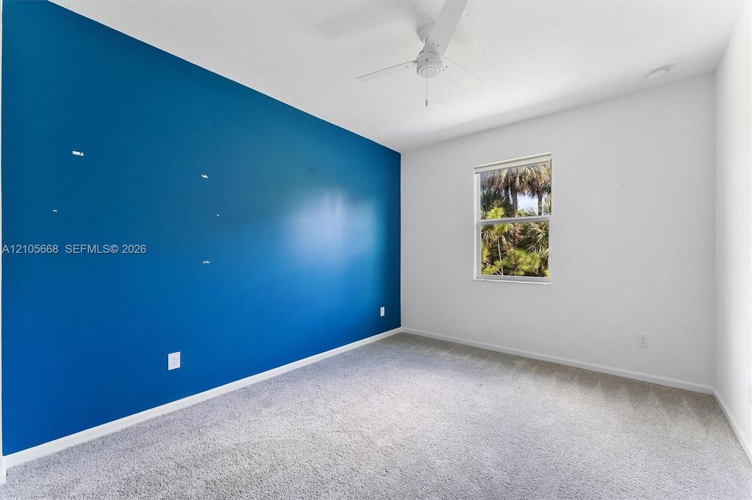 Interior space featuring a vibrant blue accent wall, neutral carpeting, a multi-blade ceiling fan, and a single window with exterior greenery