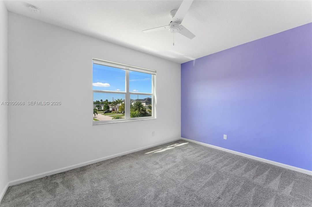 Neutral-toned wall interior featuring a vibrant accent wall, gray carpeting, a multi-blade ceiling fan, and a single-pane window