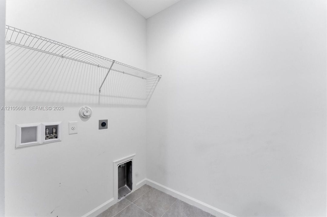 Utility area featuring white walls, gray tile flooring, installed wire shelving, and recessed plumbing connections