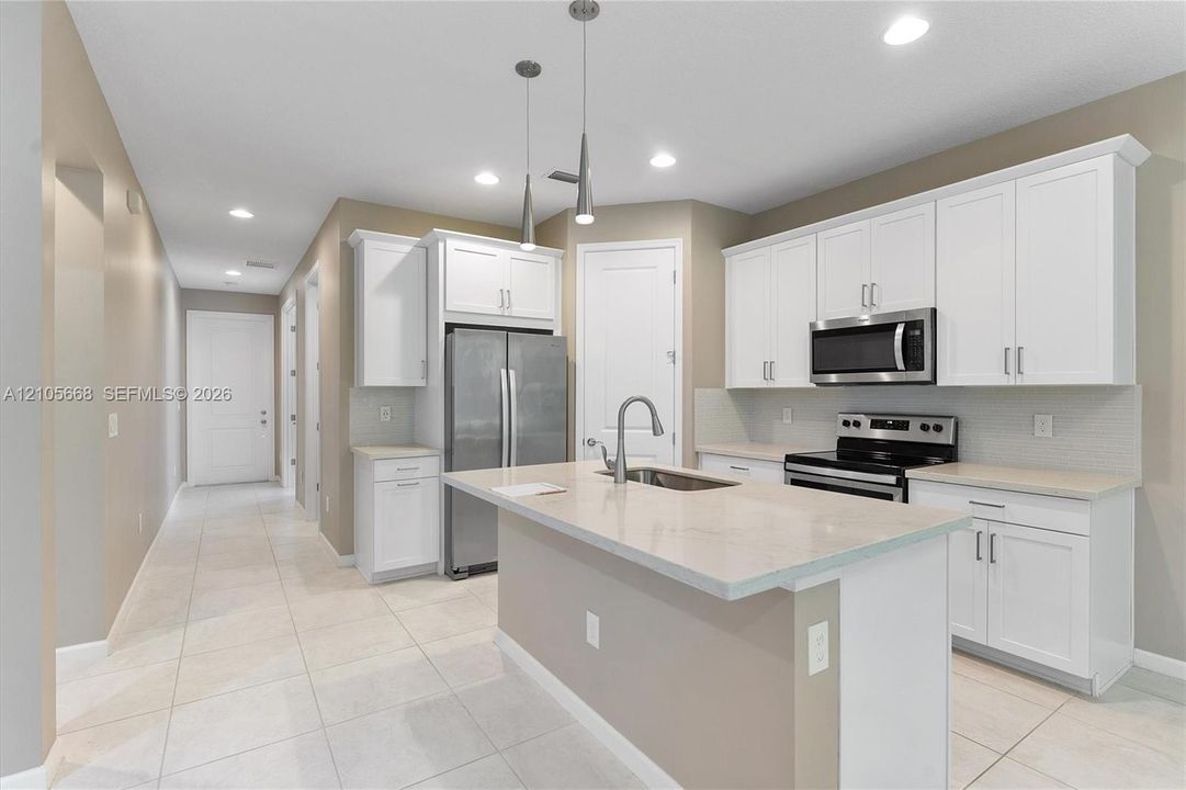 Kitchen featuring white shaker-style cabinetry, a central island with a stainless steel sink, light-toned stone-finish countertops, stainless steel appliances, and pendant lighting