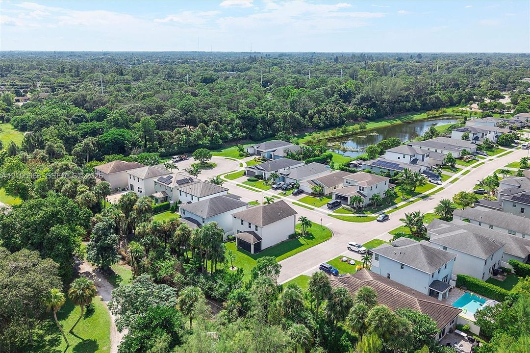 Aerial perspective of a suburban neighborhood featuring multi-story residential structures, integrated paved roadways, mature wooded canopy, and a central retention pond