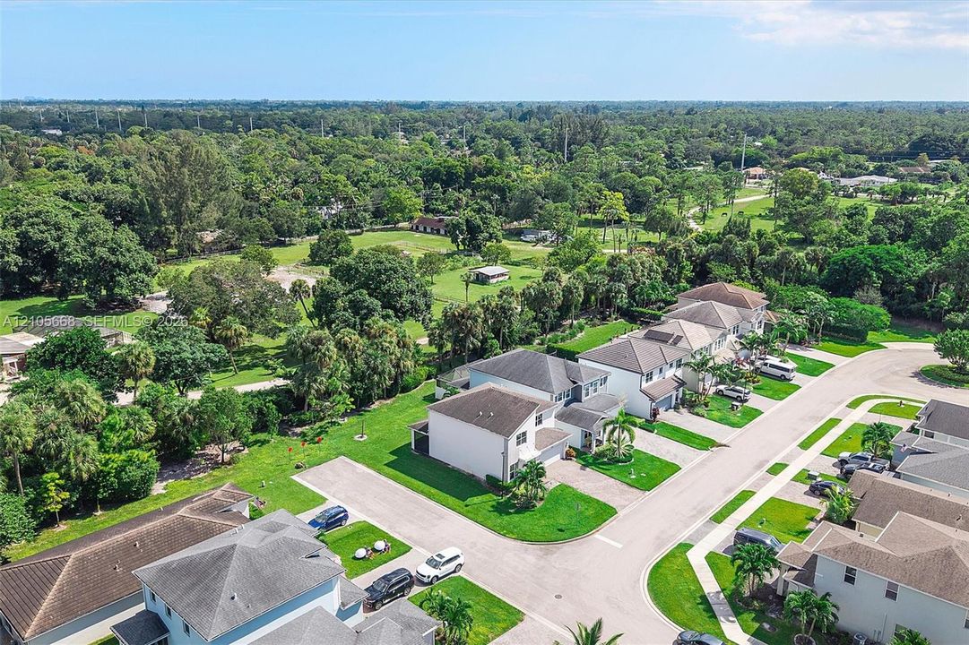 Aerial view featuring residential properties with shingled roofs, integrated multi-car garages, manicured lawns, and expansive surrounding green space with mature trees
