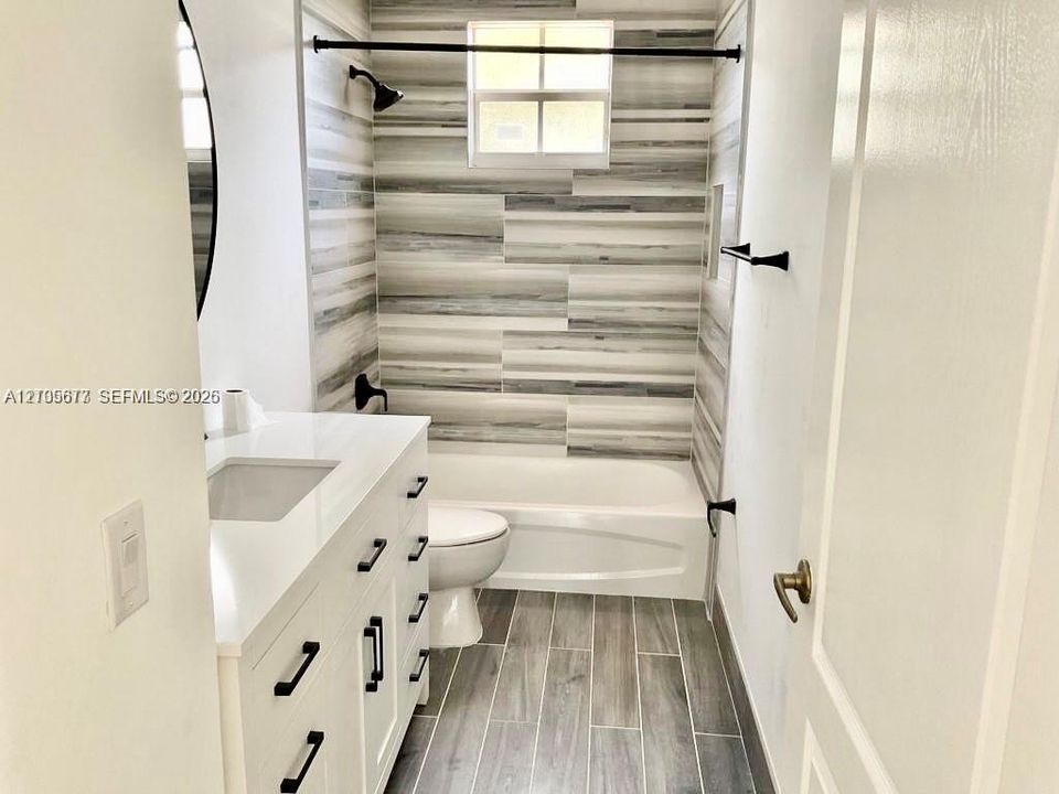 Bathroom featuring a white vanity with a white solid-surface countertop, matte black hardware, horizontal multi-tonal wall tile in the tub enclosure, and wood-finish tile flooring