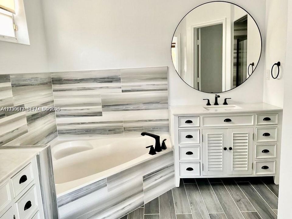 White vanity with louvered cabinet doors, quartz countertop, corner soaking tub with matte black faucet, and horizontal wall tile surround