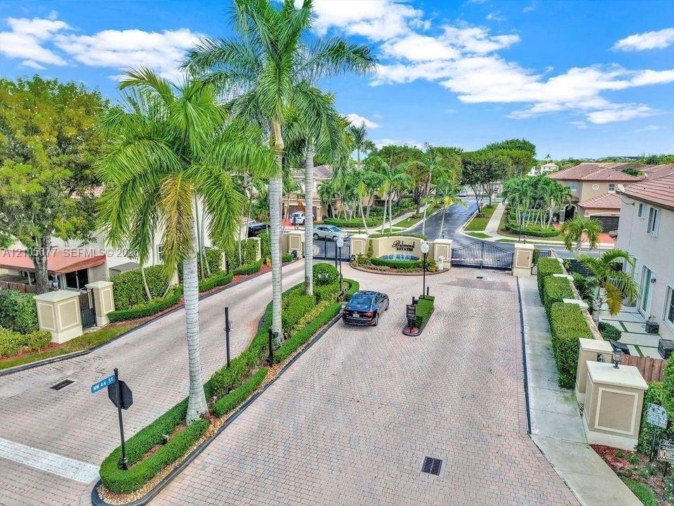 Gated community entrance featuring paver-stone driveways, ornamental stone pillars, professional landscaping with mature palm trees, and structural security gate