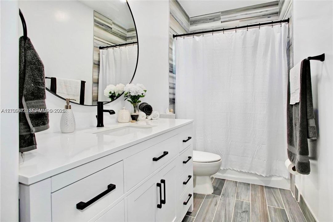 White vanity with solid-surface countertop and matte black hardware, integrated sink, circular mirror, and wood-finish tile flooring