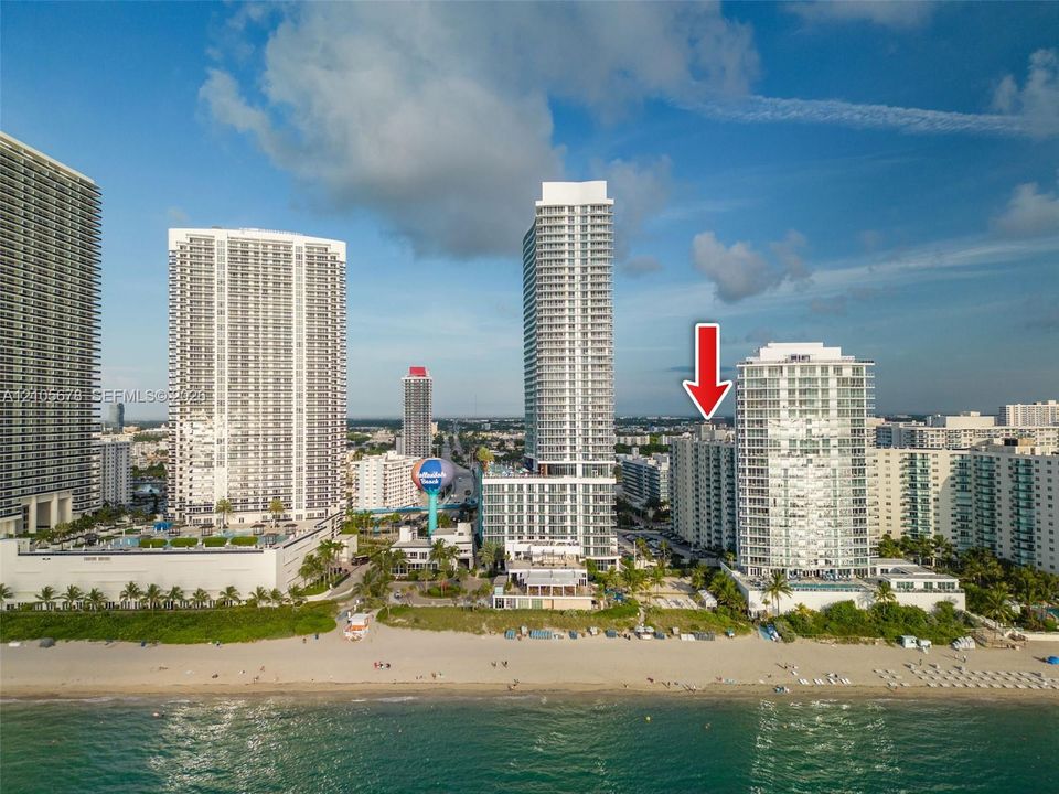 Aerial perspective of a high-rise residential building situated along a coastal shoreline, featuring direct beach access and proximity to regional urban centers