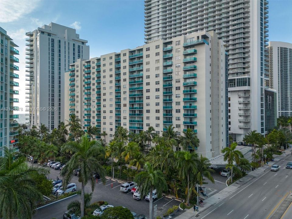 High-rise building exterior featuring private balconies with glass railings, paved surface parking, and mature palm landscaping