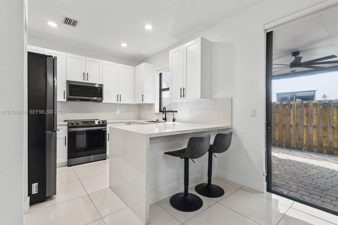 Kitchen featuring white shaker-style cabinetry, quartz countertops with an integrated peninsula, black stainless steel appliances, and large-format tile flooring with access to a paver patio