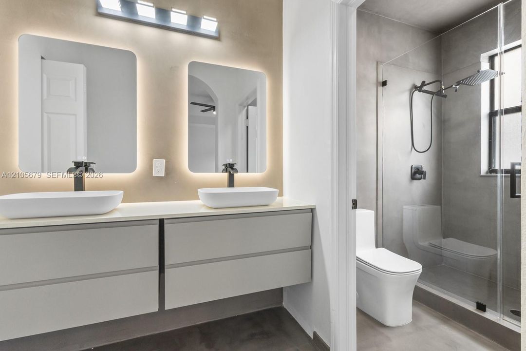 Floating double vanity with dual vessel sinks, matte black fixtures, backlit mirrors, and a glass-enclosed walk-in shower