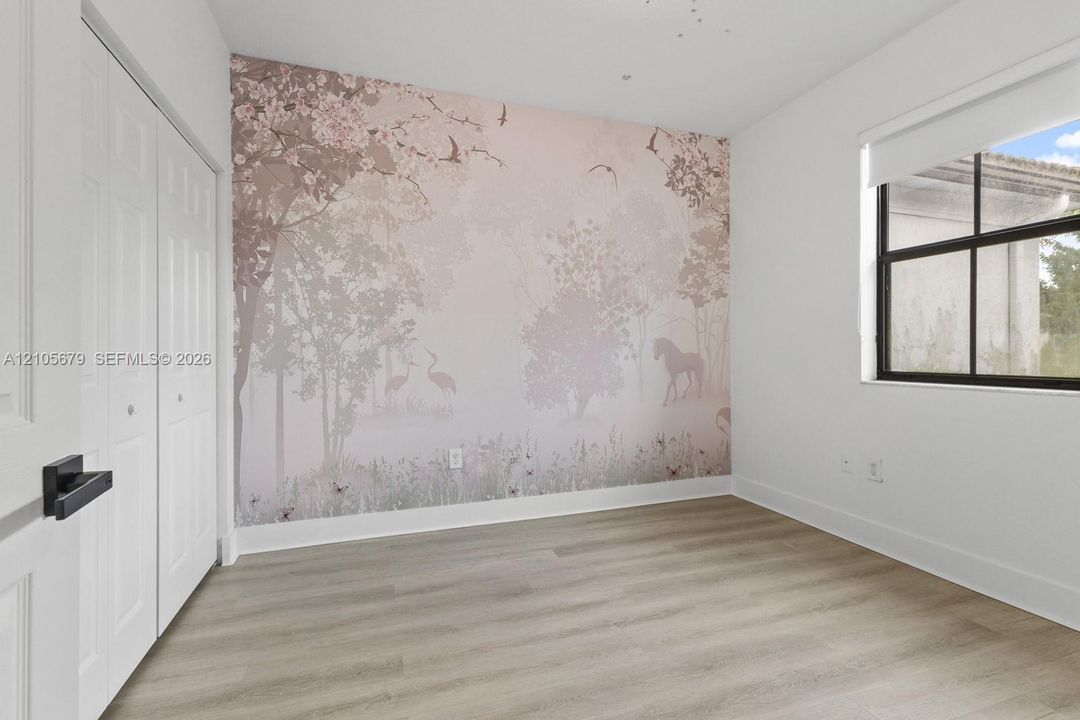 Room featuring wood-finish flooring, a decorative mural wall covering, and a single-pane window with a roller shade, complemented by white bi-fold closet doors with matte black hardware