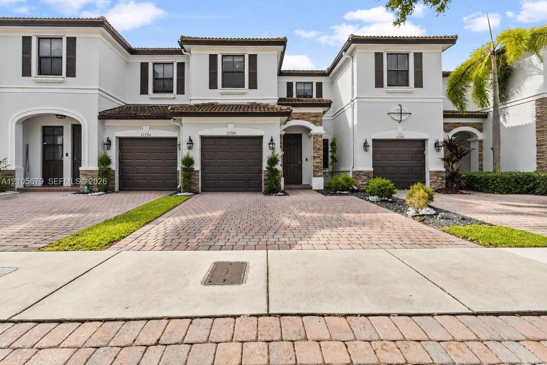 Mediterranean-inspired townhome exterior featuring multi-toned stone masonry accents, paver driveways, decorative shutters, and tile roofing