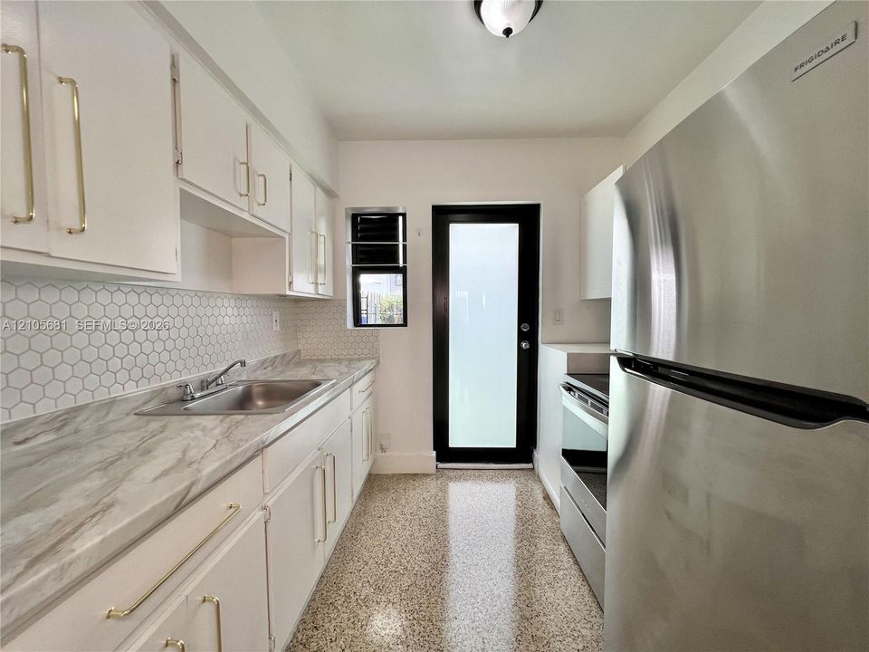 Kitchen featuring white cabinetry with gold-tone hardware, stone-finish countertops, a hexagonal tile backsplash, stainless steel refrigerator, and terrazzo flooring