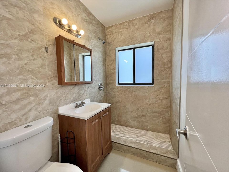 Full-height stone-finish wall tiling, walk-in shower with mosaic tile flooring, wood-finish vanity cabinet with integrated sink, and triple-bulb wall-mounted light fixture