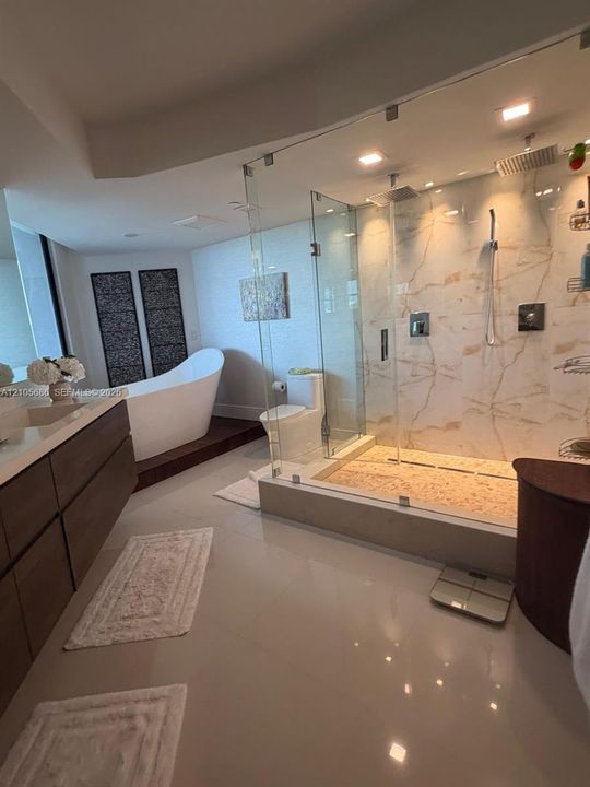 Bathroom featuring a walk-in glass-enclosed shower with large-format stone-patterned wall tile, pebble-texture shower flooring, a freestanding soaking tub set on a wood-finish platform, and a dual-drawer floating vanity with a light-toned solid surface countertop