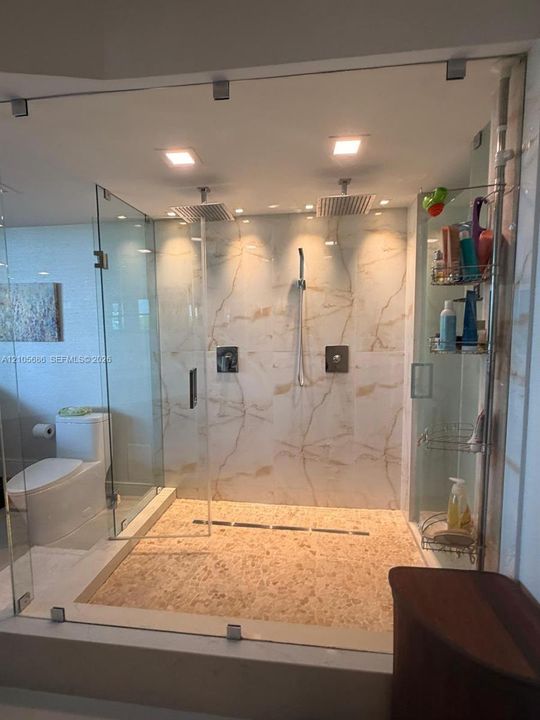 Walk-in shower featuring a frameless glass enclosure, dual rain-style showerheads, hand-held sprayer, large-format stone-look wall tile, and a textured stone mosaic floor
