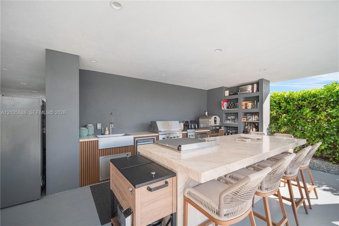 Covered outdoor kitchen featuring a stone-topped island, stainless steel farmhouse sink, built-in stainless steel grill, and custom wood-slat cabinetry with integrated shelving
