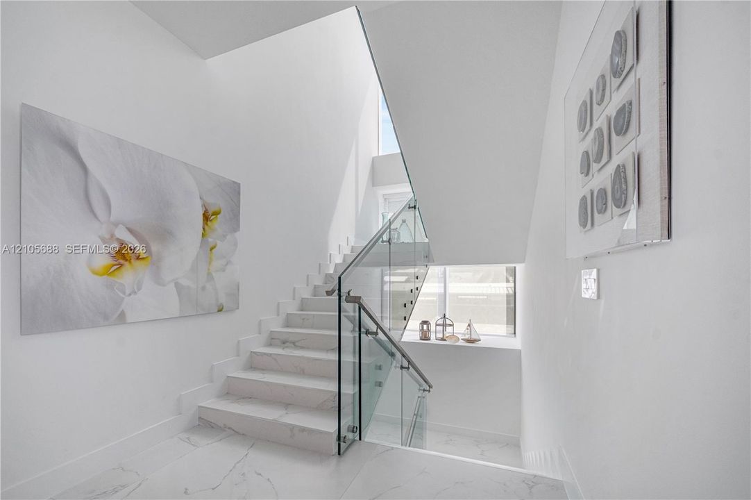 Staircase featuring white stone-finish steps, glass balustrade, metal handrails, and a multi-level window design