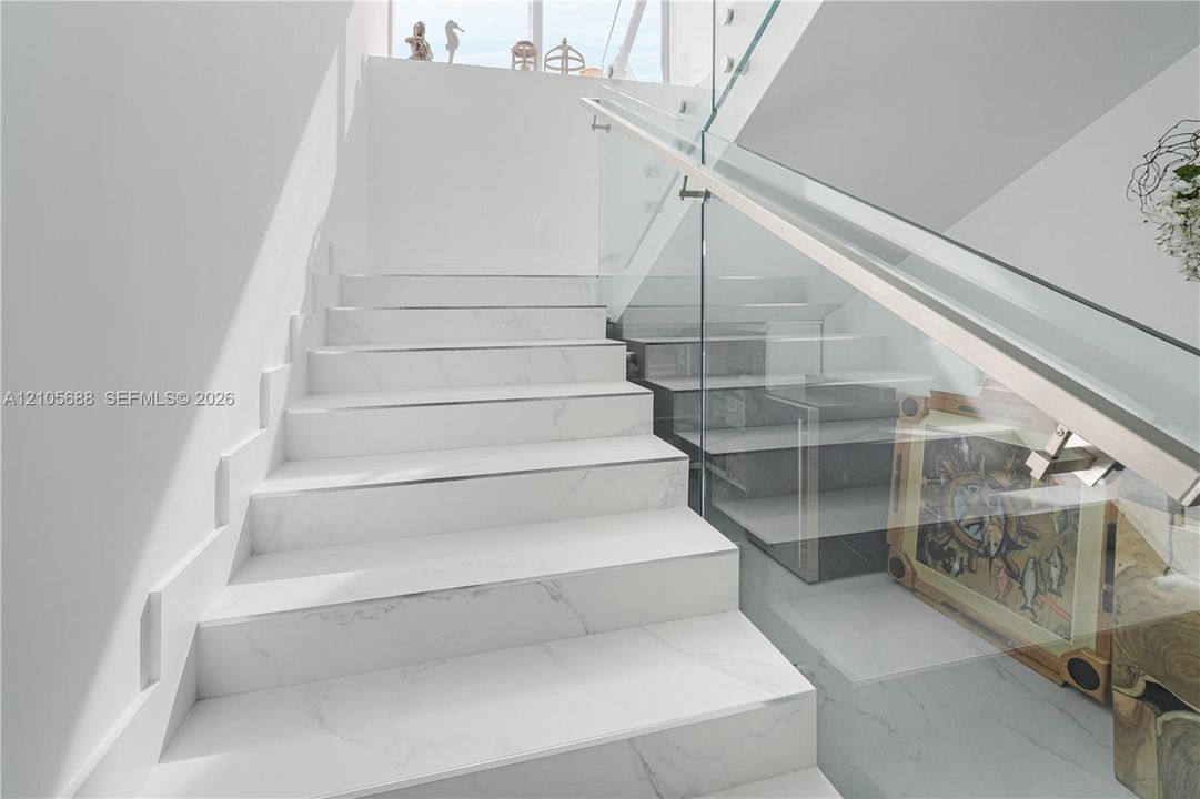 Staircase featuring white stone-finish treads, glass railing panels, and a polished metal handrail