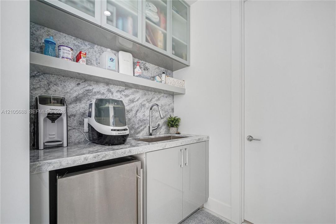 Wet bar area featuring high-gloss cabinetry, stone-patterned countertops and backsplash, integrated stainless steel under-counter refrigeration, an undermount sink with a gooseneck faucet, and upper glass-front cabinetry