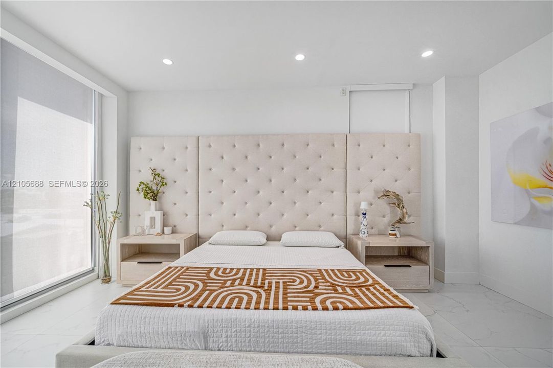 Large-format tile flooring, recessed ceiling lighting, floor-to-ceiling glass window panel, and built-in bedside cabinetry featuring integrated drawer pulls