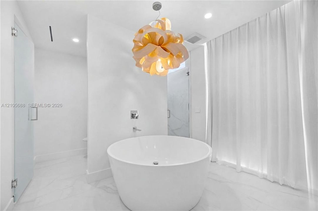 Freestanding circular soaking tub with a wall-mounted faucet, sculptural pendant light fixture, and stone-finish tile flooring