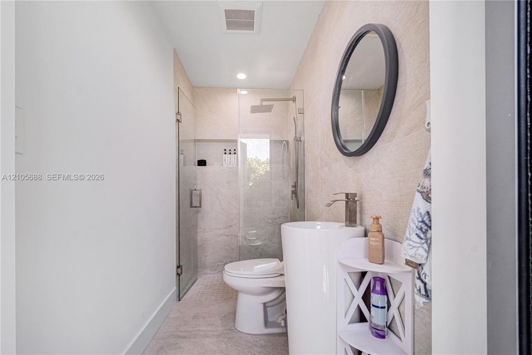 Pedestal sink with metallic faucet, round mirror, glass-enclosed shower, and textured stone-finish wall tile