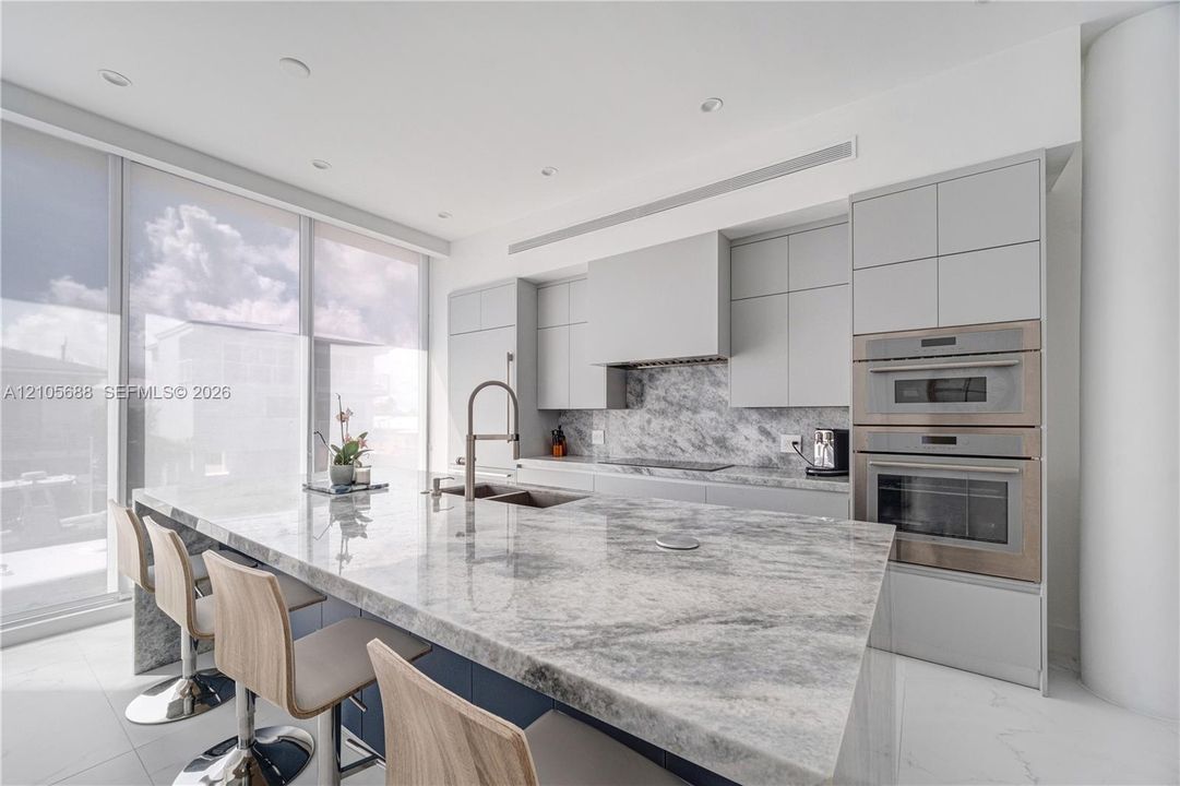 Modern kitchen featuring floor-to-ceiling windows, a large stone-surfaced waterfall island, matching stone slab backsplash, flat-panel cabinetry, and integrated stainless steel double wall ovens