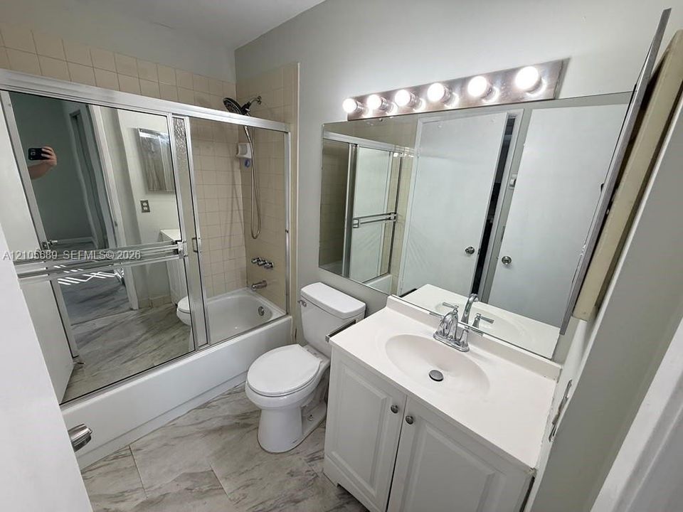 Bathroom featuring a vanity with a solid-surface countertop, full-width vanity mirror, tile-surround tub-shower combination with sliding glass doors, and light-tone stone-finish flooring