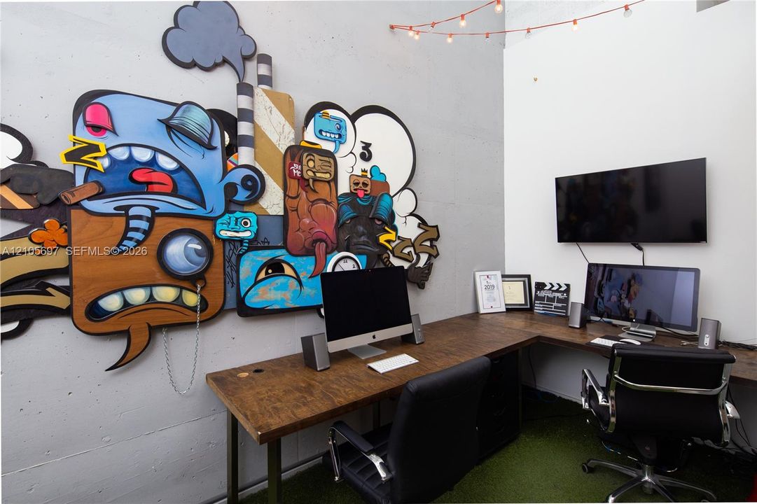 Exposed concrete walls featuring a large-scale, multi-dimensional wall-mounted art installation, integrated L-shaped built-in desk with a wood-finish surface, and artificial turf flooring