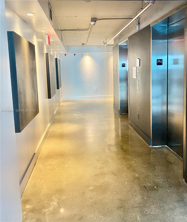 Industrial-style hallway featuring polished concrete flooring, exposed overhead conduit, recessed lighting, and stainless steel elevator doors